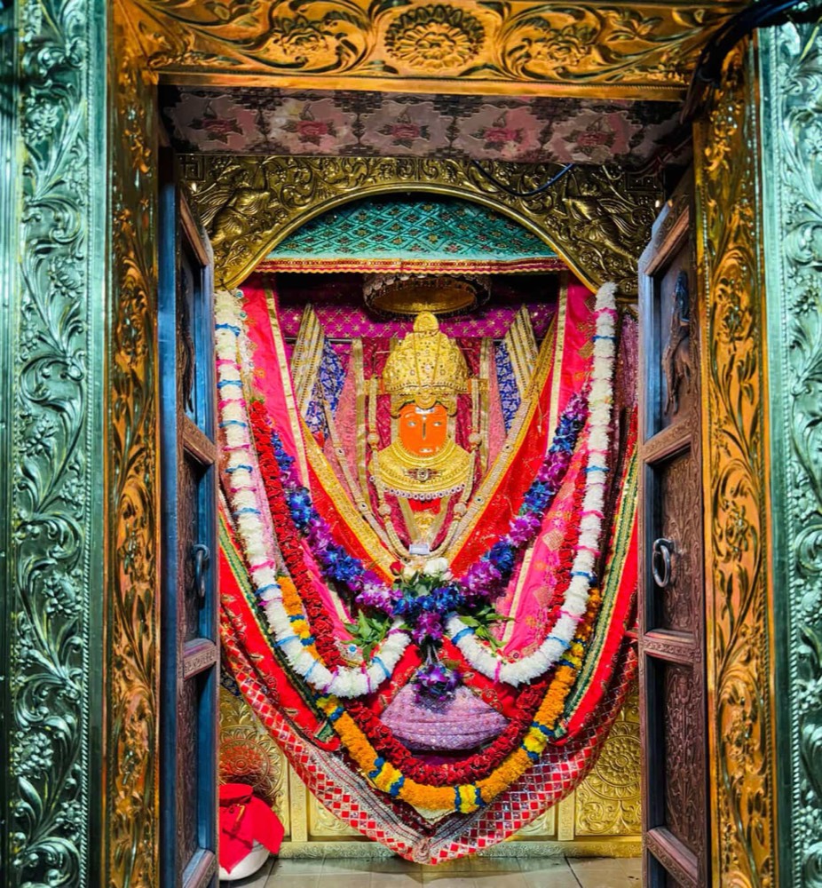 Full garbhagriha view of Jeen Mata between ornate golden doors, adorned with red and pink silks and long white, blue and red flower garlands