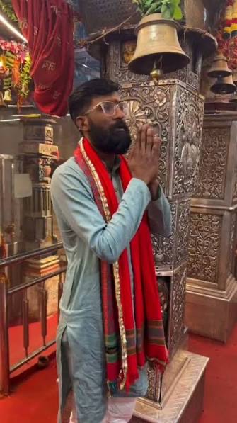 A devotee wearing a red and gold dupatta stands with folded hands in prayer beside an ornate silver pillar and temple bells inside the Jeen Mata temple
