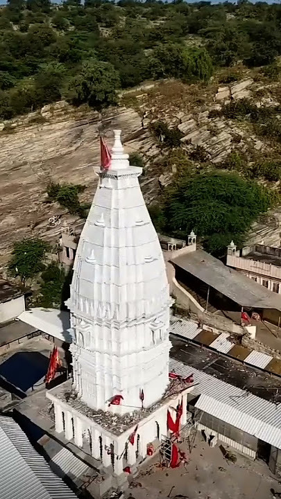 Aerial view of the white, carved shikhar of the Jeen Mata temple with red flags, set against the rocky, tree-dotted Aravalli hillside